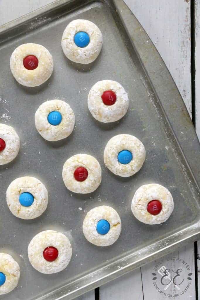 Thumbprint cookies rolled in powdered sugar and pressed with red and blue M&Ms are evenly placed on a baking pan.