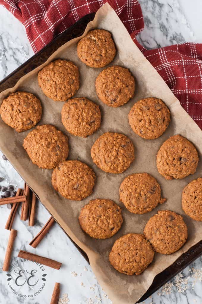 Flat lay of Oatmeal Pumpkin Chocolate Chip Cookies on parchment paper on a baking sheet, surrounded by a red towel and cinnamon sticks, on a marble counter top.