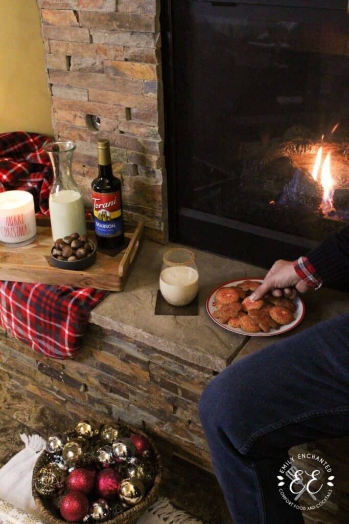 A fireplace mantle with a plate of cookies, holiday drink, and a tray with bowl of chestnuts, Torani syrup, pitcher of eggnog, and a lit Christmas candle