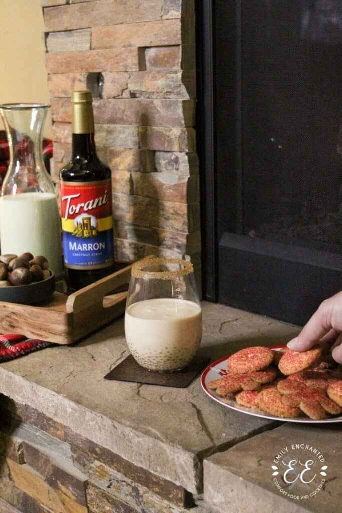 A fireplace mantle with a plate of cookies, holiday drink, and a tray with bowl of chestnuts, Torani syrup, and pitcher of eggnog