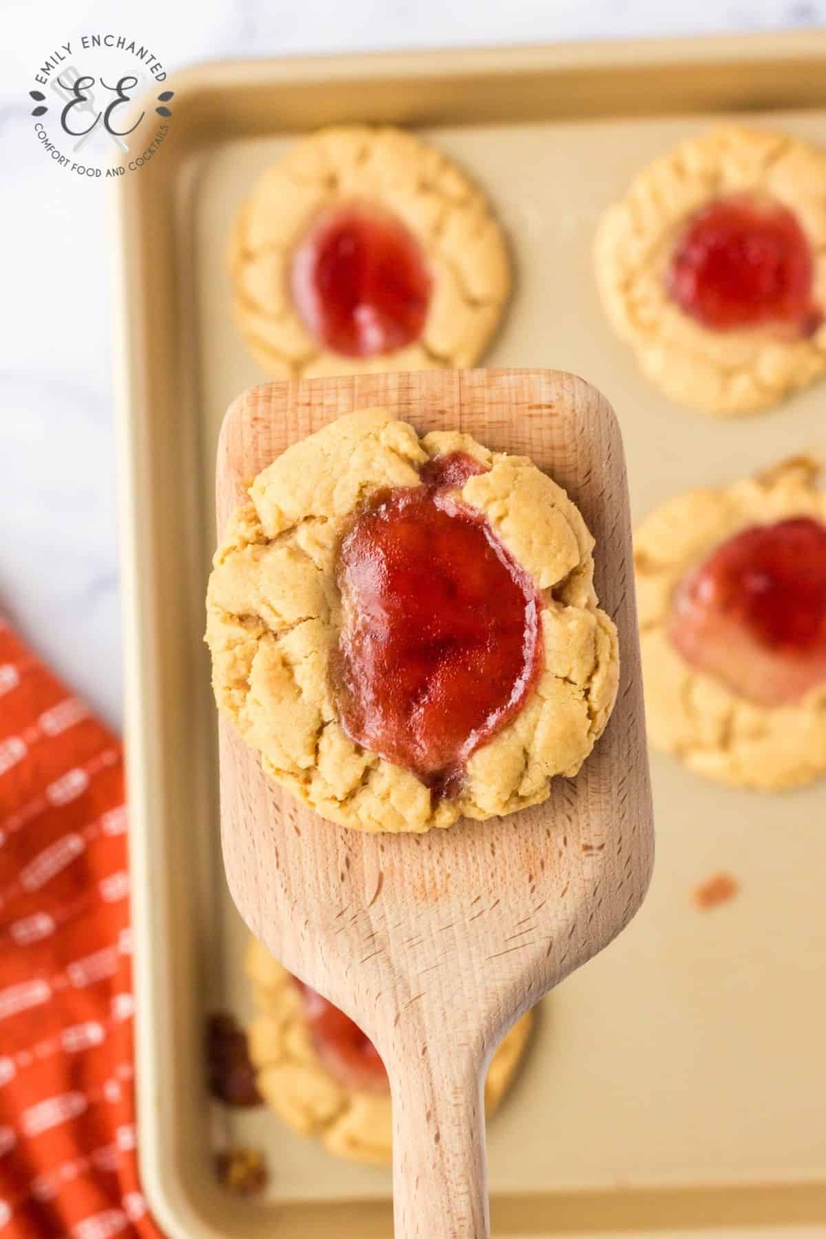 Close up of peanut butter cookie with jelly on a wooden spatula with tray of cookies in background